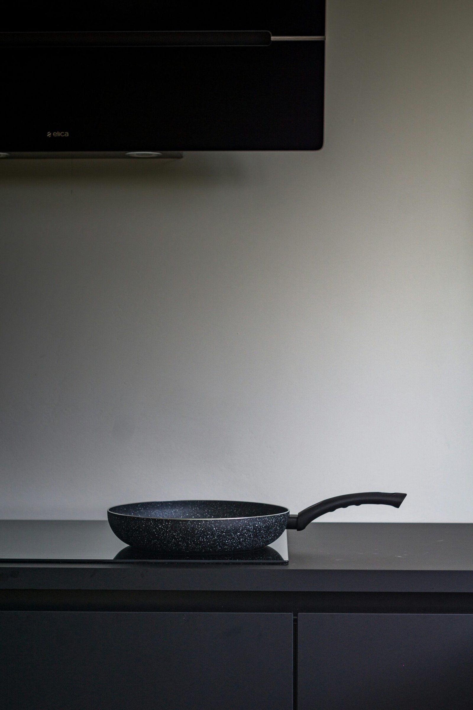 A black non-stick frying pan with a handle resting on a modern black stovetop in a minimalist kitchen.